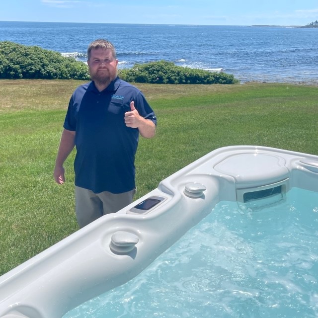 A water care technician giving a thumbs up next to a hot tub outside in front of the ocean.