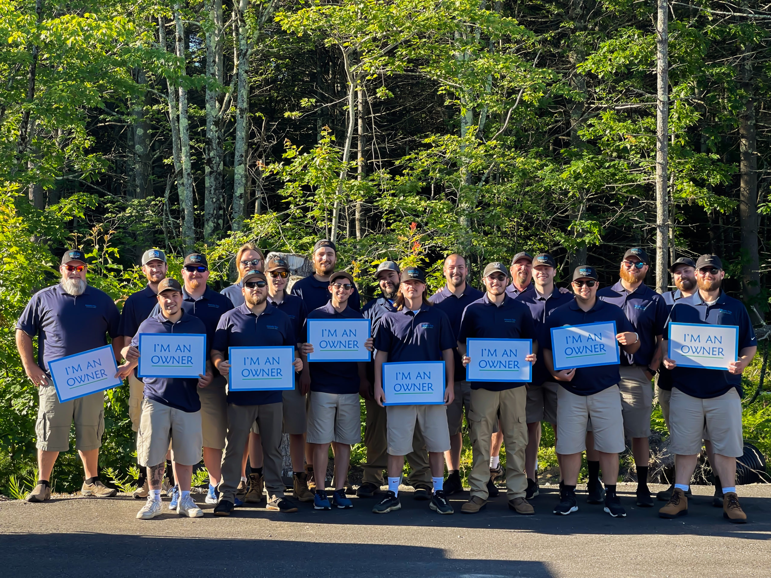 Group of employees standing outside in front of trees holding 'I'm an owner' signs.