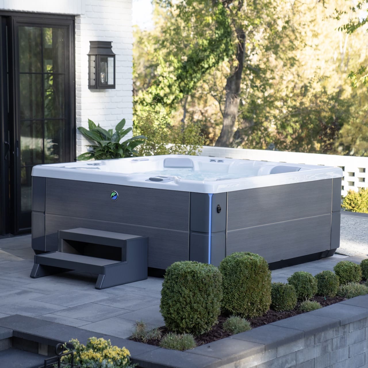 Highlife aria hot tub in alpine white and charcoal sitting on a grey patio with green trees and shrubs surrounding the patio