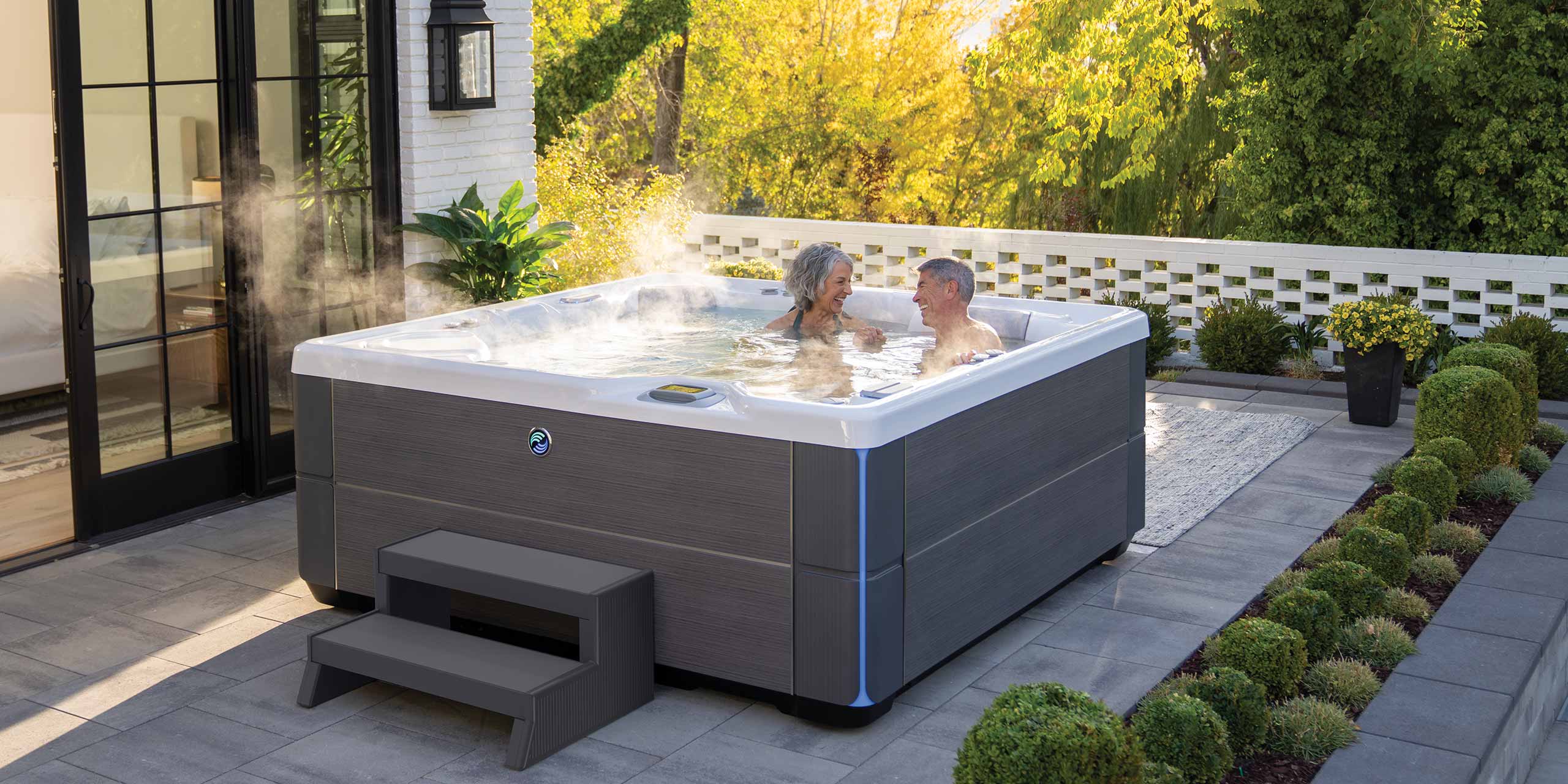Older man and woman sitting in the highlife aria hot tub, laughing in the water. The hot tub is Alpine white & Charcoal and they are on a patio with green shrubs surrounding the hot tub.